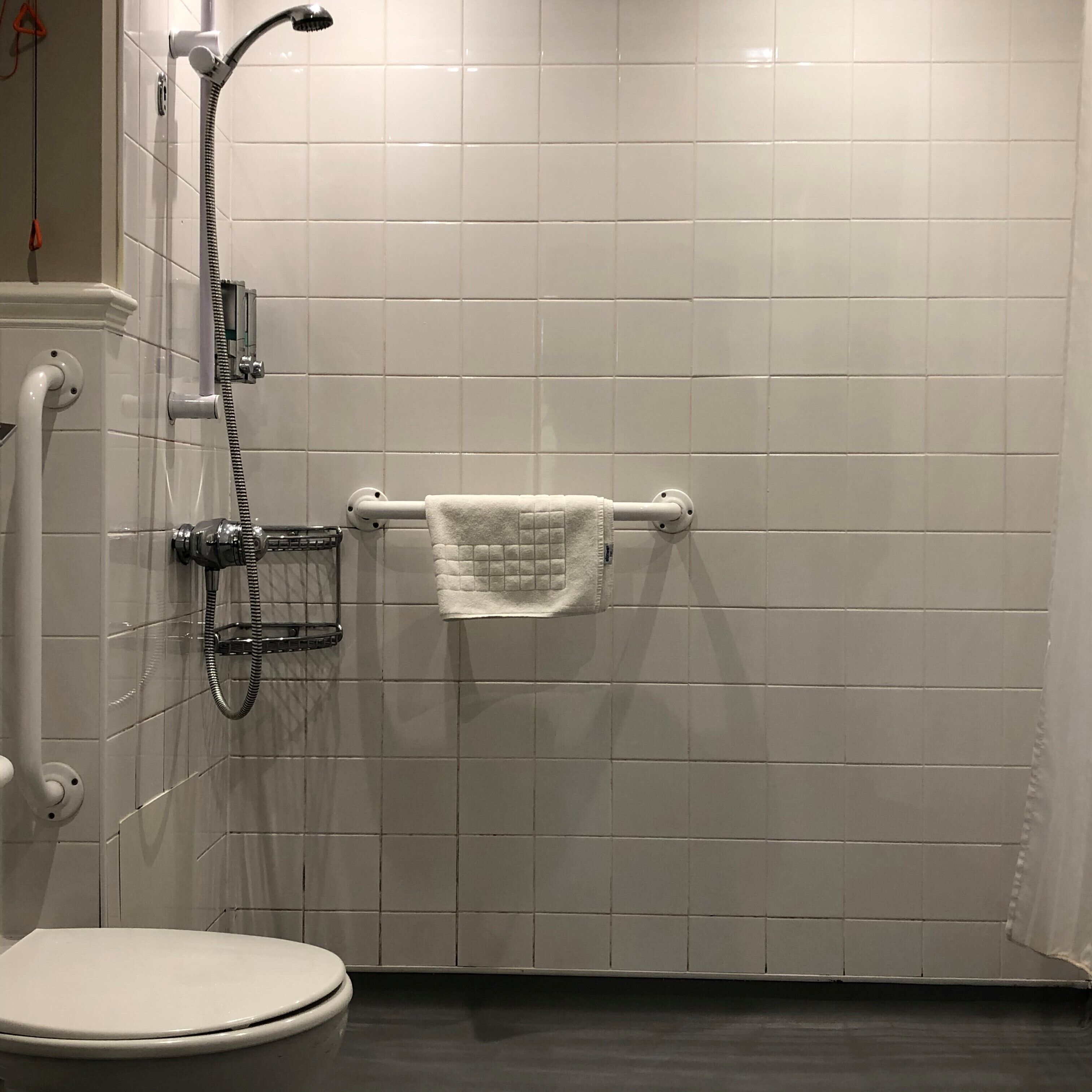 A bathroom with white tiled walls, a white toilet, a shower with a handheld showerhead, and a towel rack with a towel.