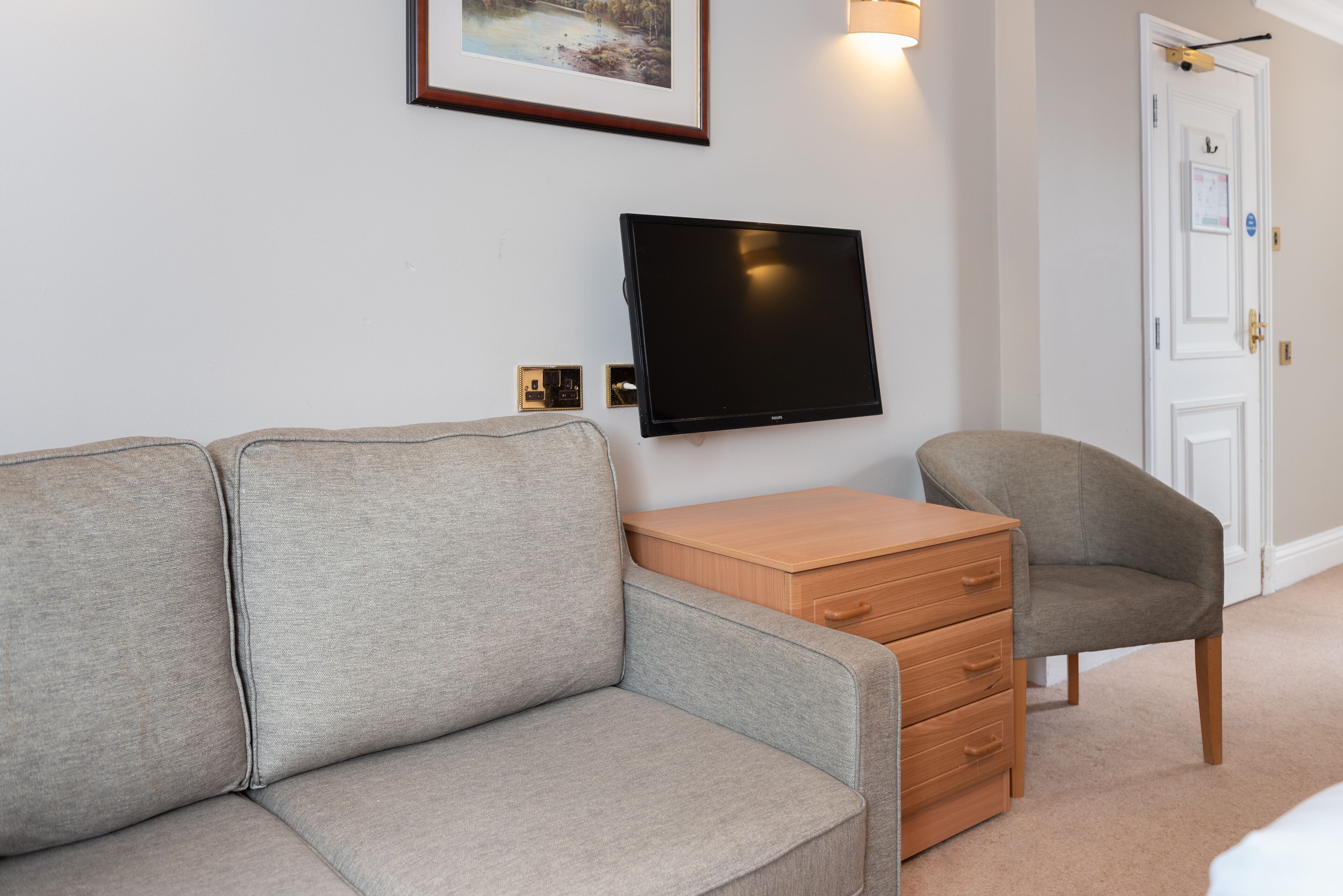A room with a grey sofa, a wooden nightstand, a television mounted on the wall, and a door with a security lock.