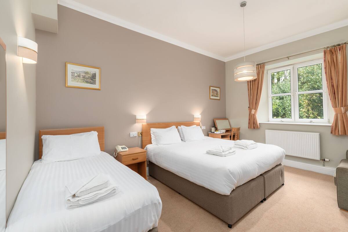 Quarto de hotel com duas camas, uma janela com cortinas, uma mesinha de cabeceira com luminária e um quadro na parede.