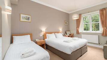 A hotel room with two beds, a window with curtains, a bedside table with a lamp, and a radiator.