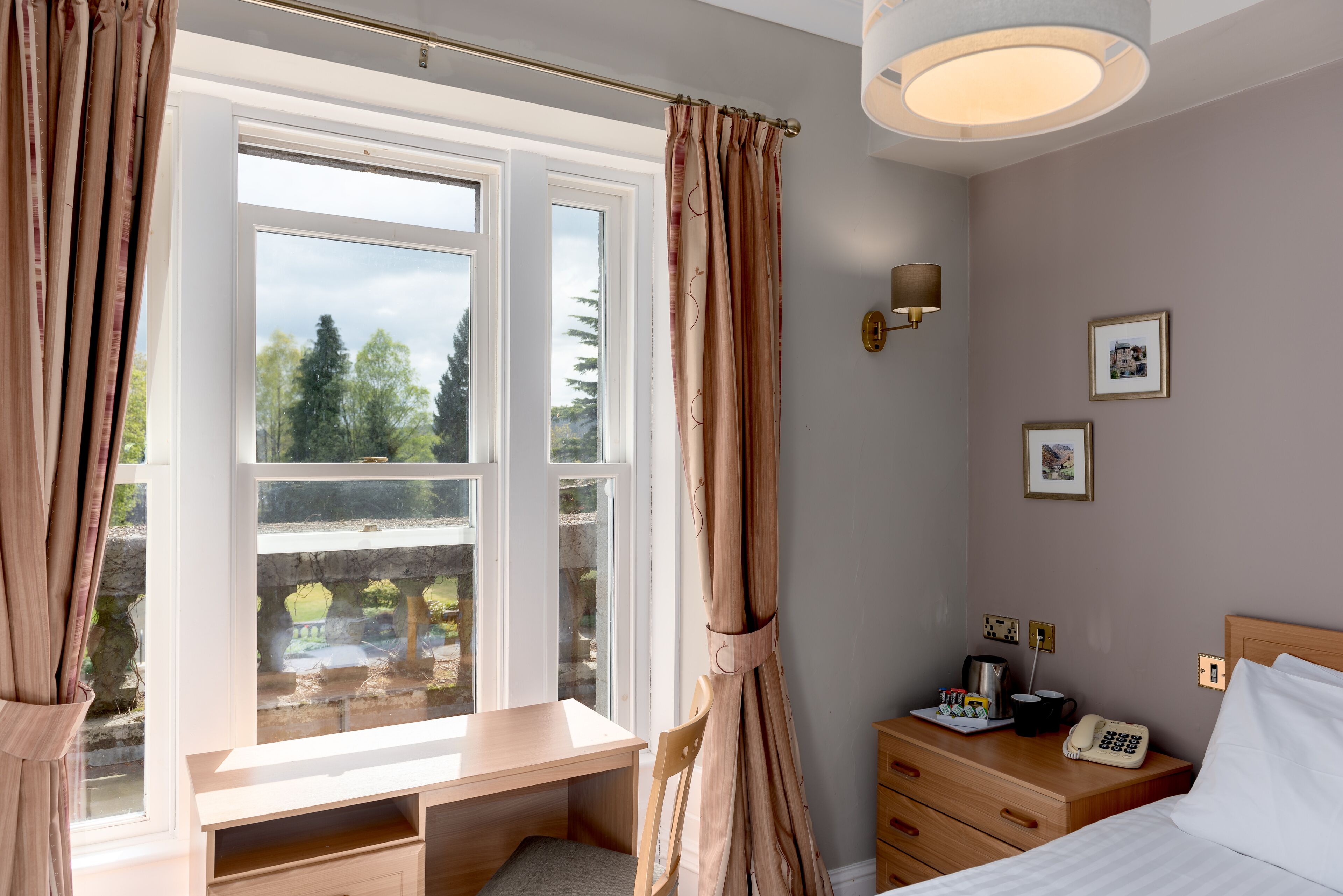 A bedroom with a desk, a bed, a bedside table with a phone, and a wall-mounted lamp.