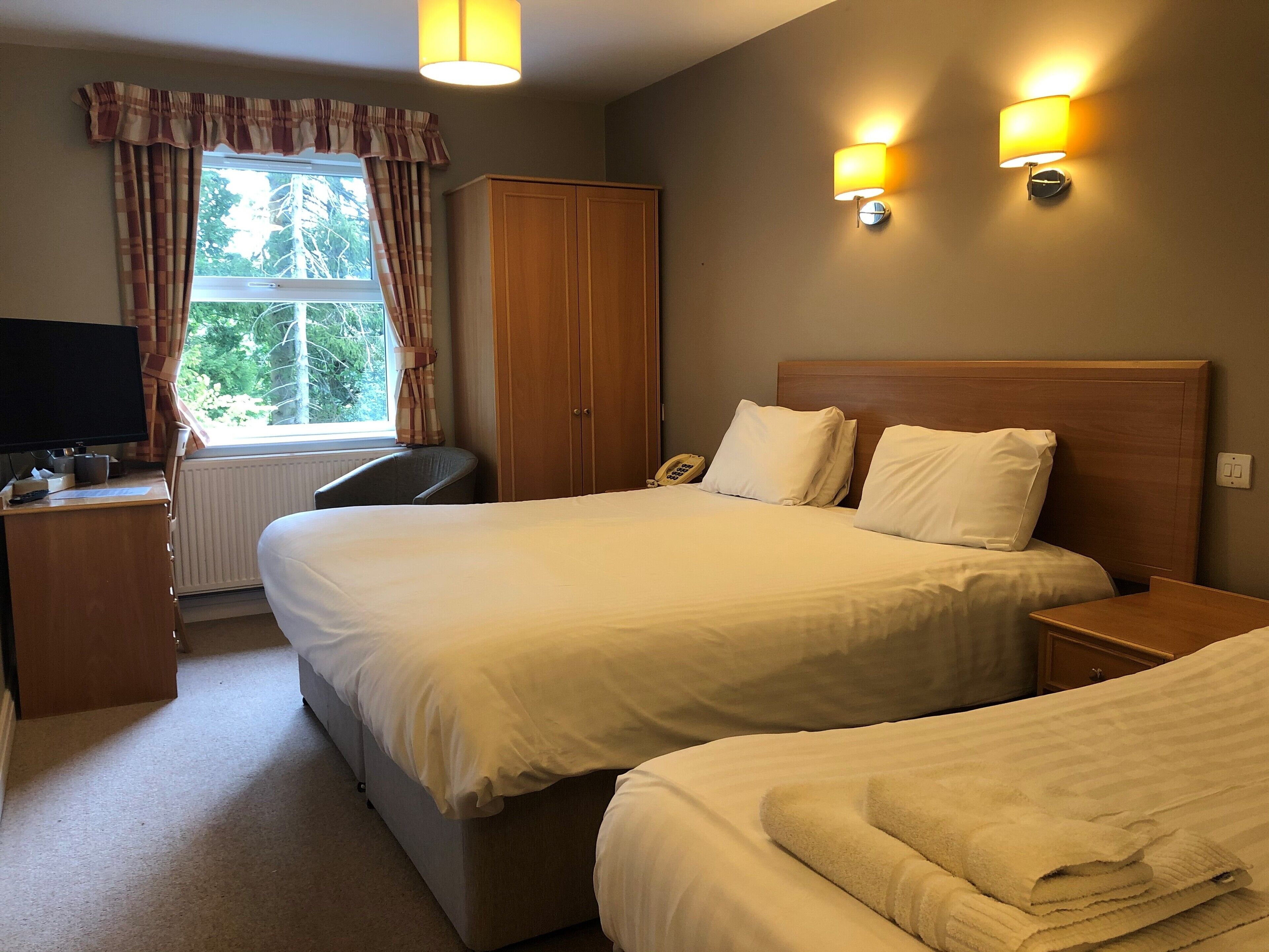 A hotel room with a large bed, a desk with a computer, a chair, and a window with curtains. There are two wall-mounted lamps and a telephone on the desk.
