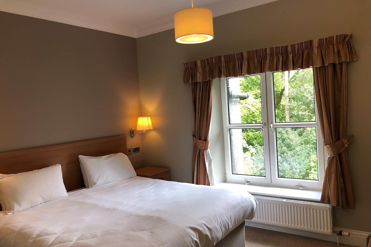 Quarto de hotel com uma cama grande, uma mesa de cabeceira, uma luminária na parede e uma janela com cortinas.