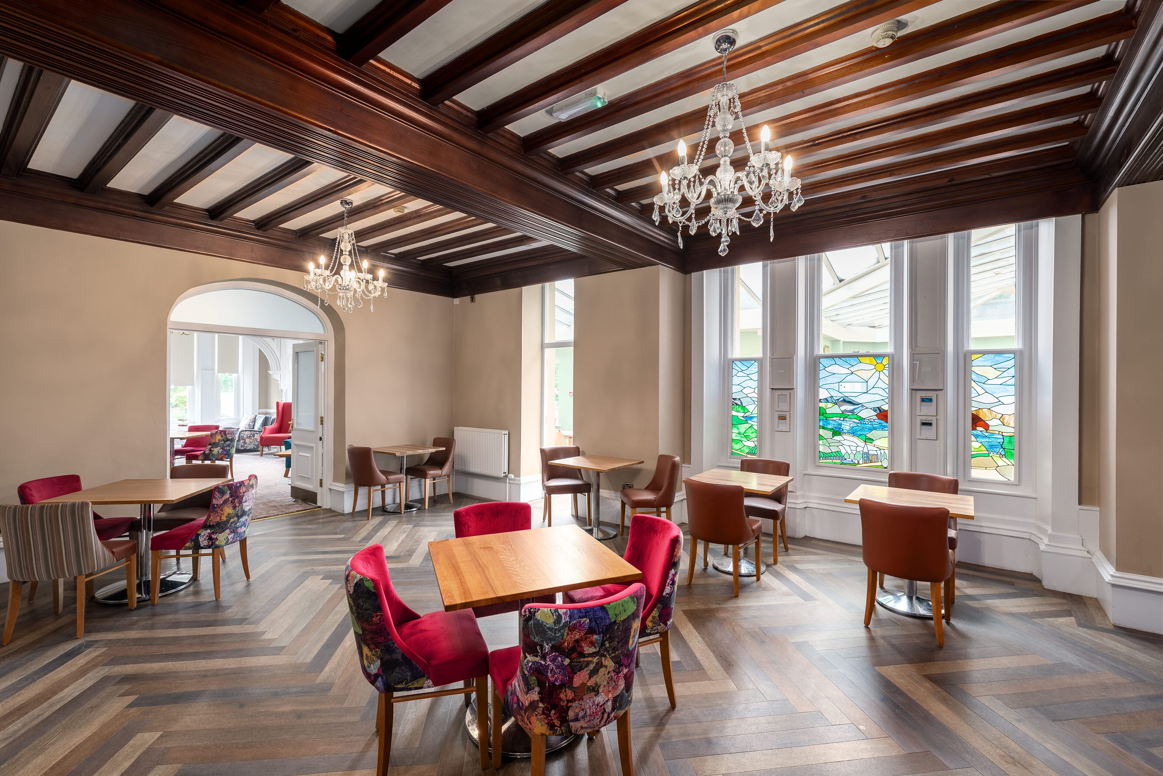 Un comedor espacioso con mesas y sillas de madera, un candelabro y ventanas amplias con vidrieras.