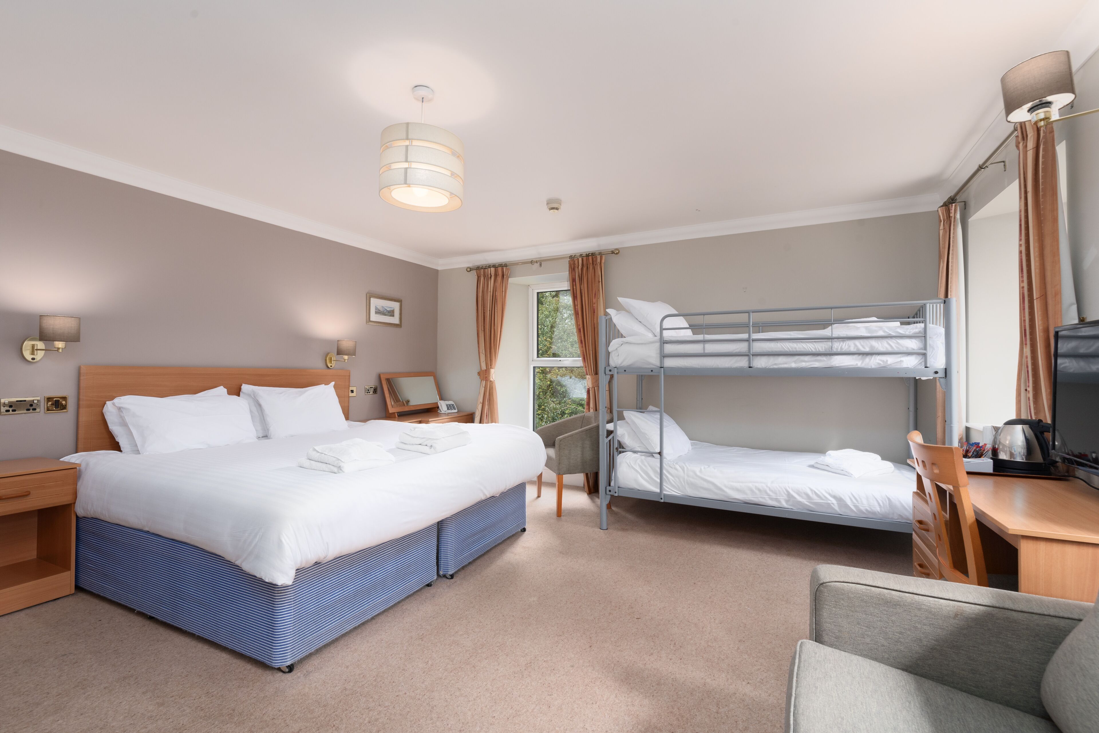 A hotel room with a large bed, a smaller bunk bed, a desk with a chair, a lamp, and a window with curtains.