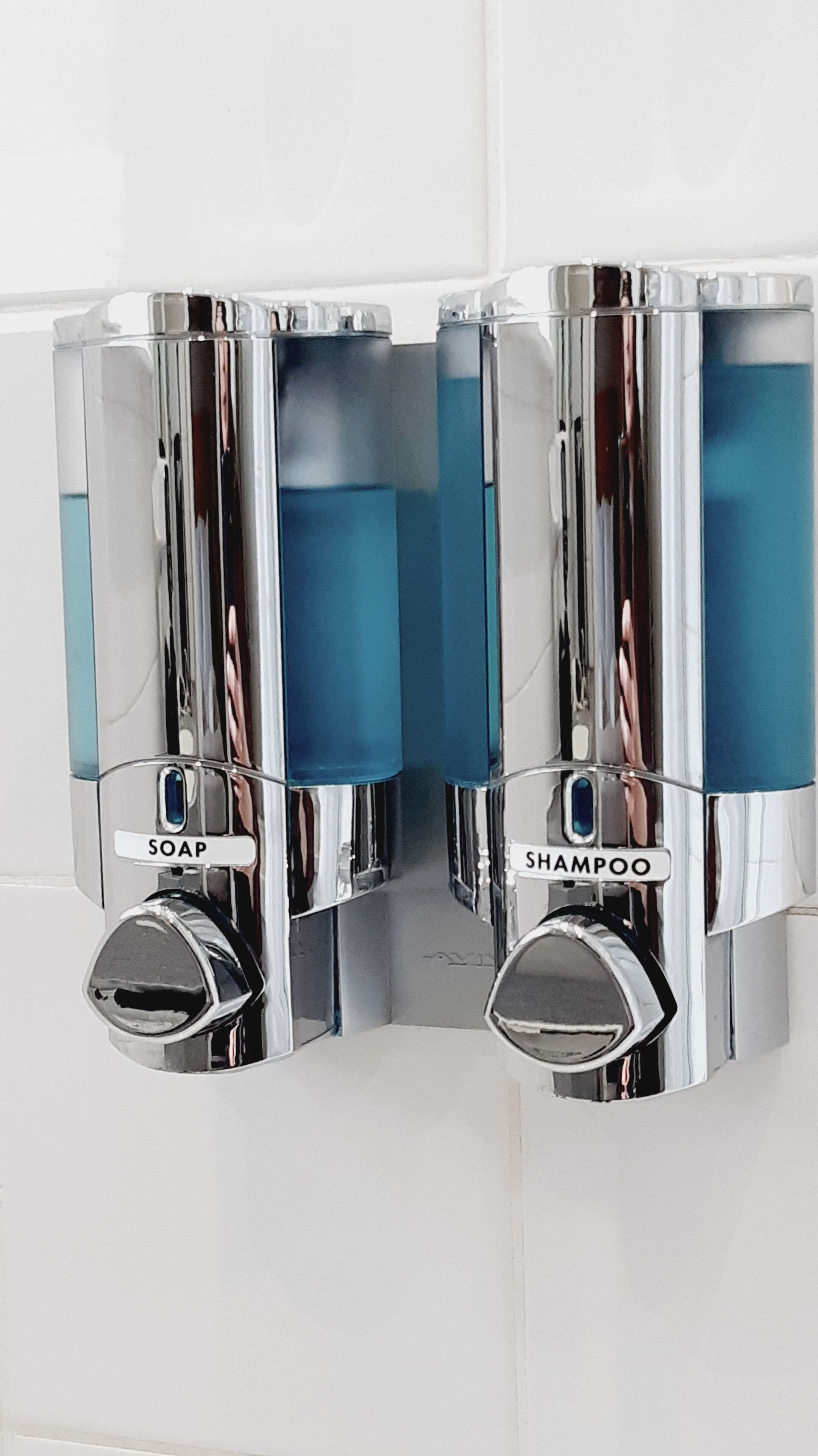 A bathroom with chrome soap and shampoo dispensers mounted on a tiled wall.