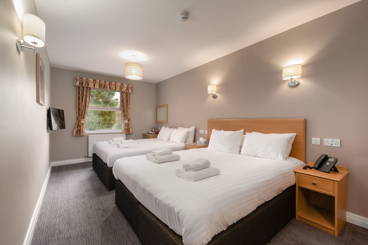 Quarto de hotel com duas camas, painel de cabeceira em madeira, luminárias laterais, telefone e uma janela com cortinas.