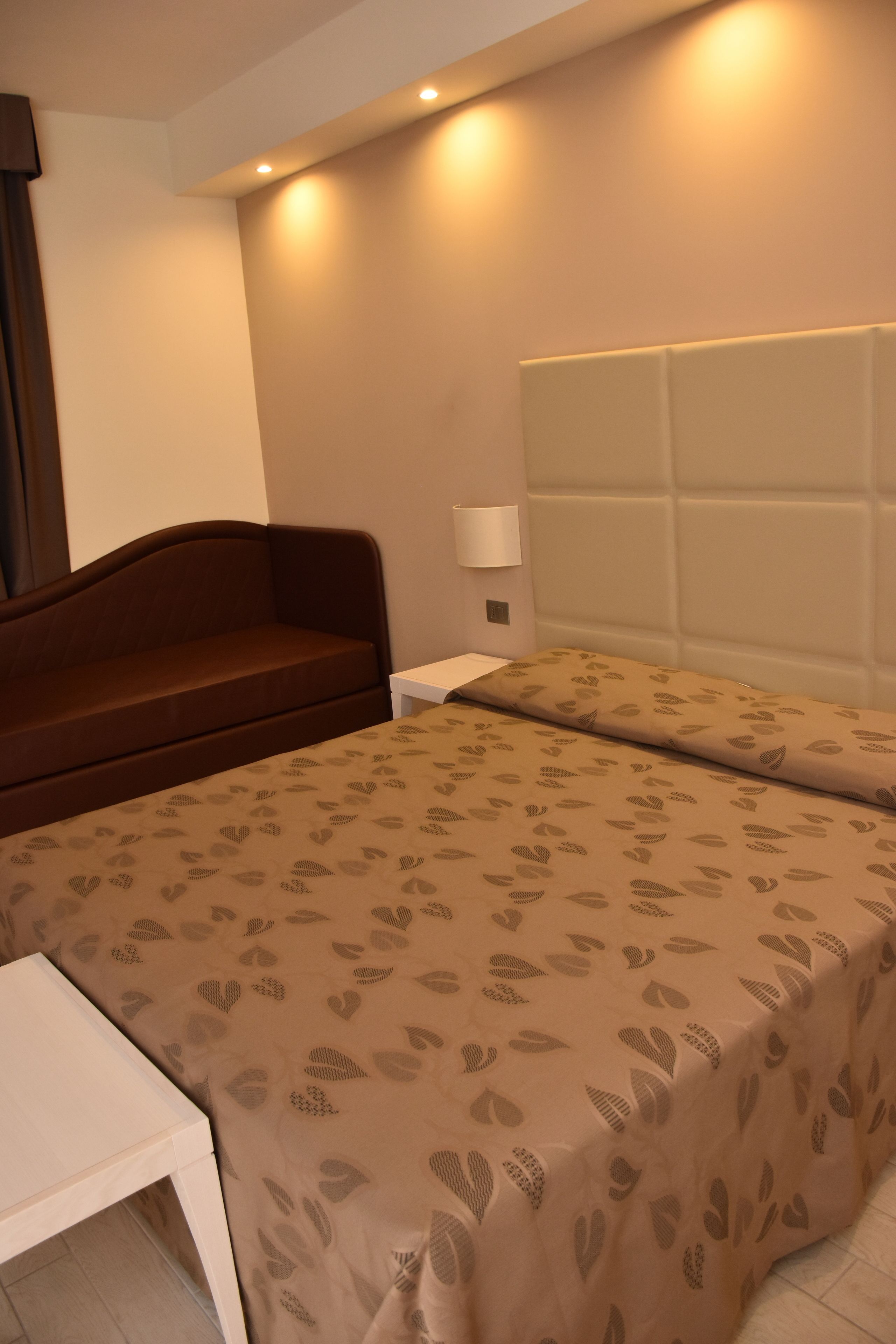 A hotel room with a bed, a bedside table, a lamp, and a brown sofa.