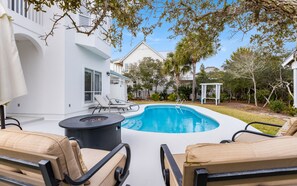 A swimming pool with lounge chairs and a fire pit, surrounded by a white building and greenery.