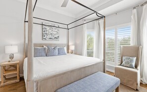 A bedroom with a four-poster bed, a bedside table with a lamp, a chair with a cushion, and a window with blinds.