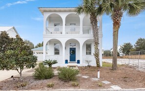 A two-story white house with a blue front door, a balcony on the second floor, and a palm tree in the front yard.
