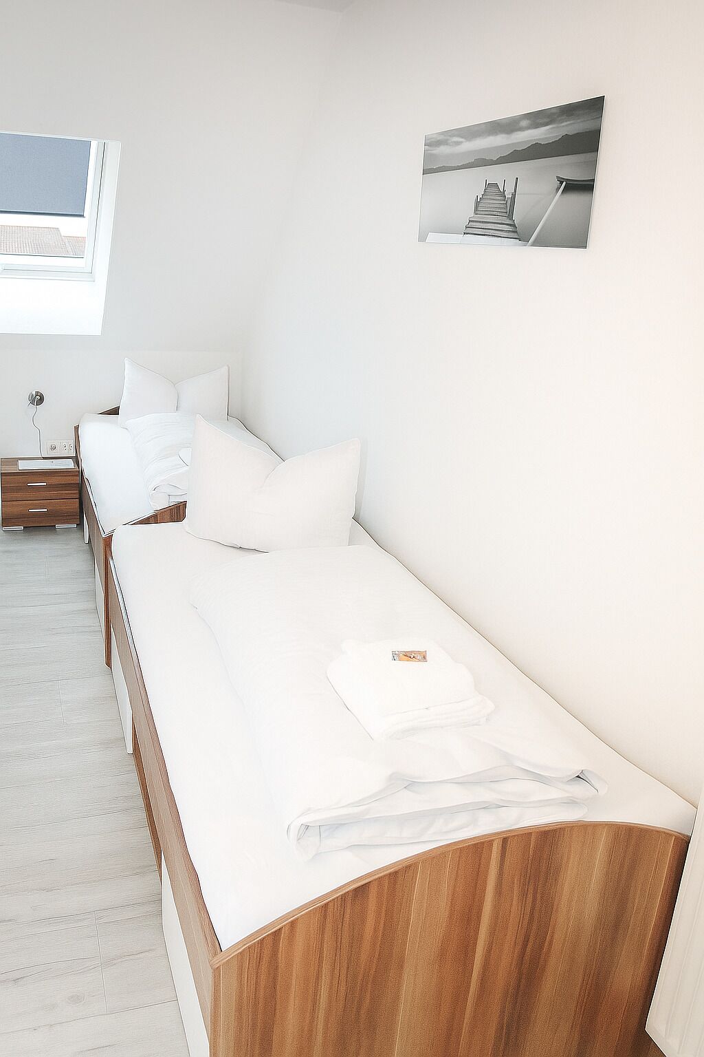 Hotellihuone, jossa on pitkä sänky, yöpöytä, ikkuna ja seinälle ripustettu taulu.