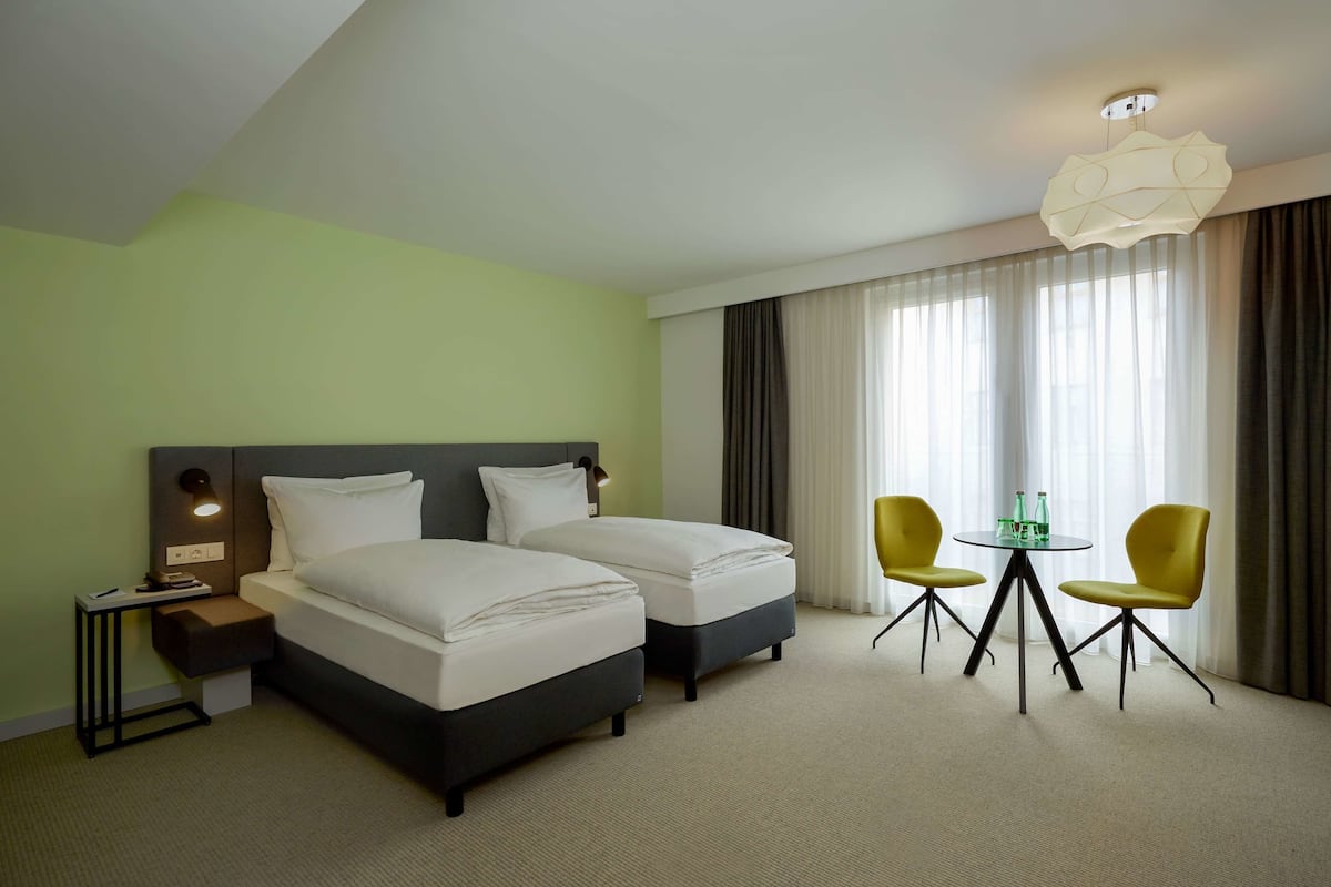 Ein Hotelzimmer mit zwei Betten, einem kleinen Tisch mit Stühlen, einem Nachttisch und einem großen Fenster mit Vorhängen.