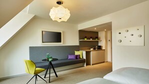 Une chambre d’hôtel moderne avec un lit en mezzanine, un canapé avec des coussins, une petite table avec une bouteille, une télévision et un élément décoratif mural.