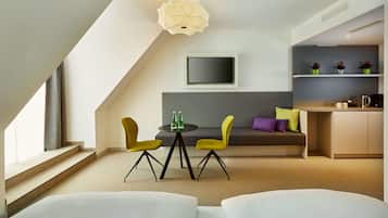 A modern room with a sofa, a dining table with chairs, a flat-screen TV, and a kitchen area with shelves.