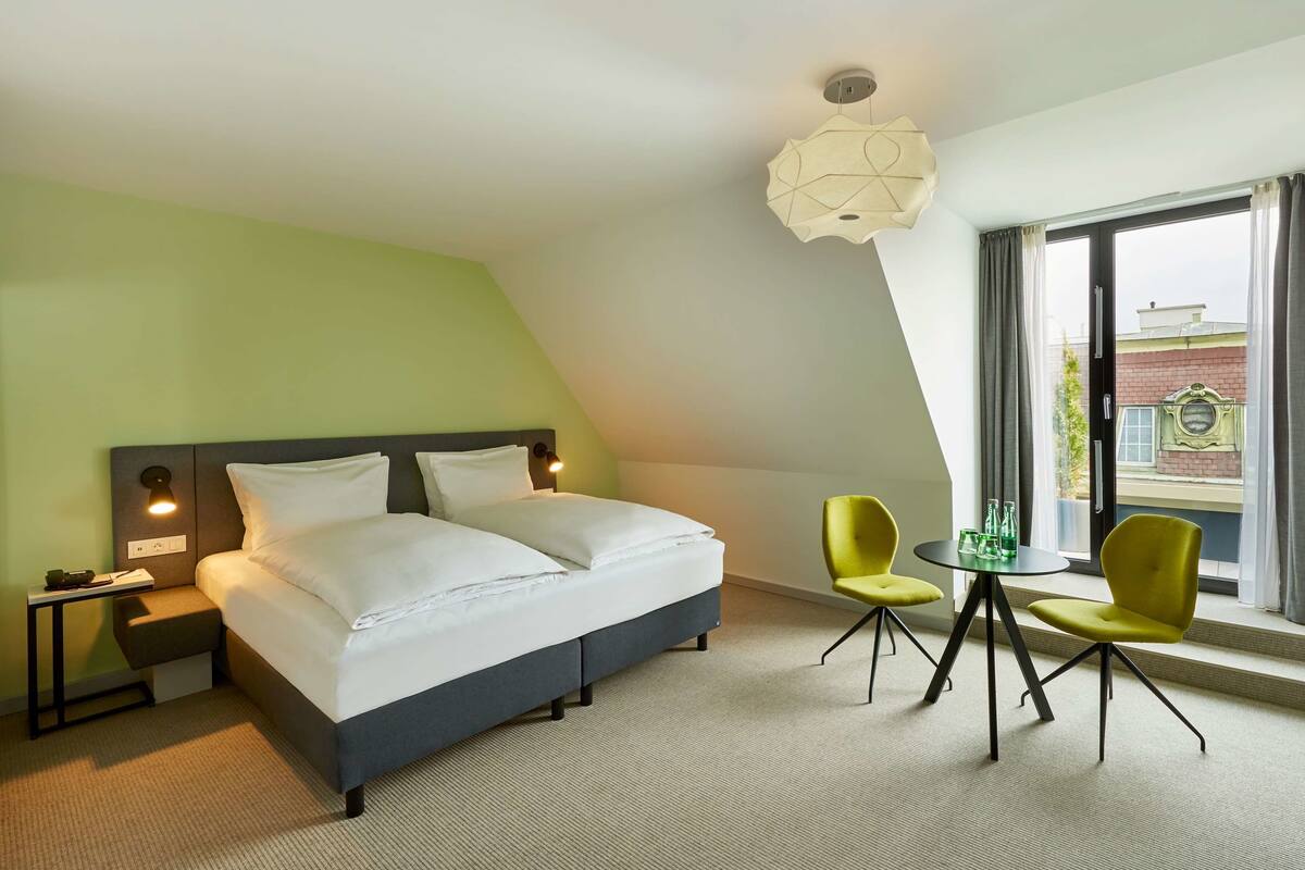 Ein Hotelzimmer mit Bett, Nachttischen, einem Schreibtisch, zwei Stühlen, einem kleinen Tisch mit Gläsern und Blick ins Freie durch ein großes Fenster.