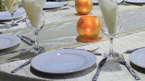Una mesa de banquete dispuesta para un evento formal, con servilletas blancas, cristalería transparente y una pantalla de proyector al fondo.