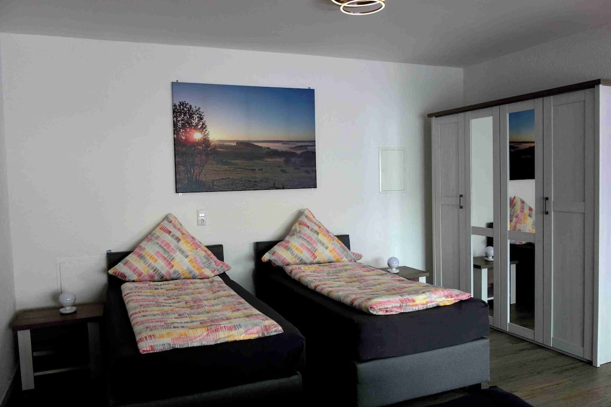 Un dormitorio con dos camas, una mesita de noche, una lámpara de pared y un ventanal grande.