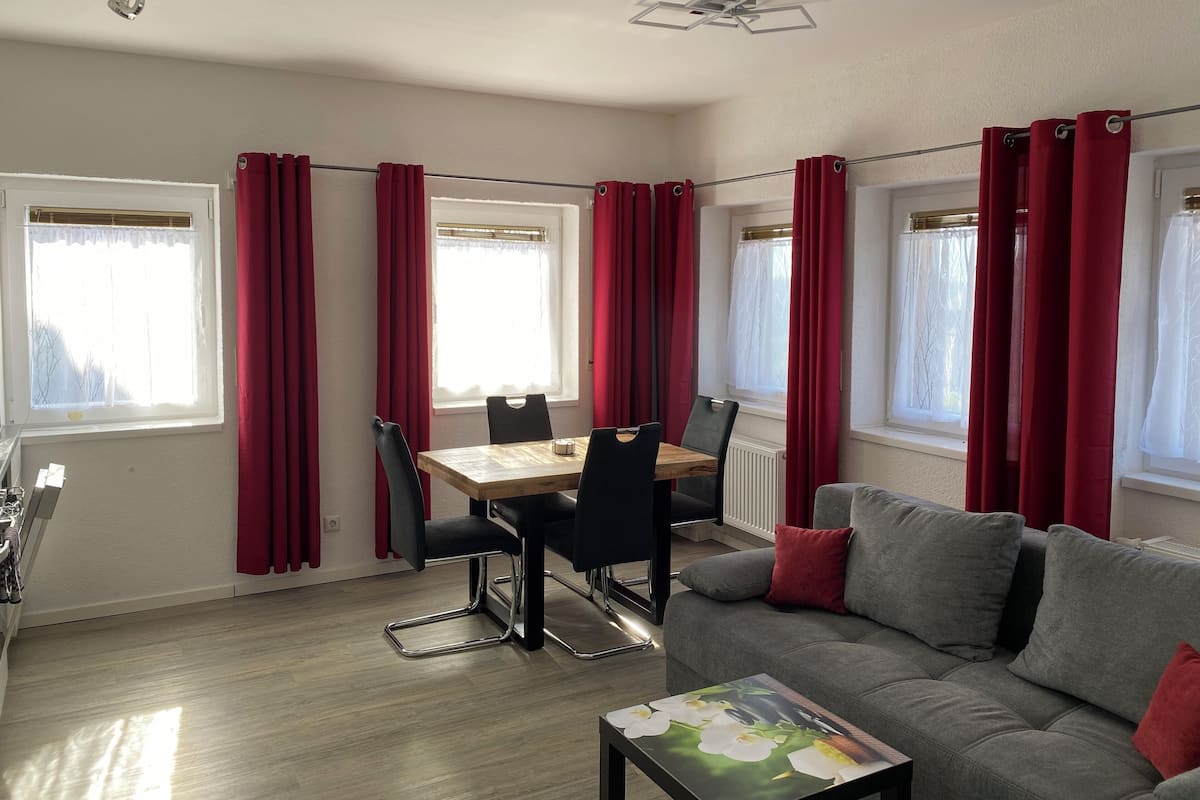 Una sala de estar moderna con un sofá gris, cortinas rojas, una mesa de comedor de madera y sillas negras.