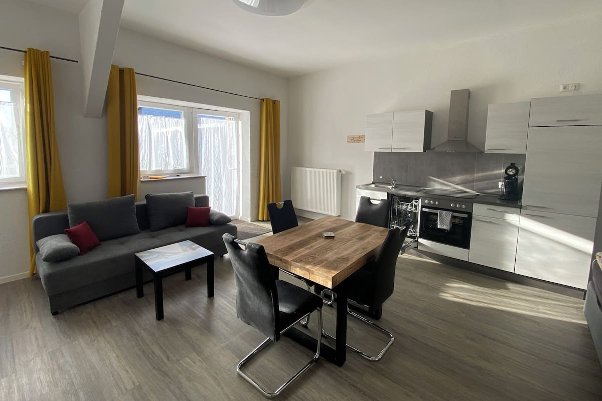 Una sala moderna con sofá, mesa de centro y mesa de comedor. Hay una cocina con estufa, fregadero y alacenas.