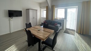 Un salon moderne avec une table à manger en bois, des chaises noires, un canapé gris agrémenté de coussins, une armoire grise et un téléviseur à écran plat fixé au mur.