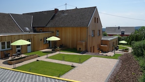 Un bâtiment moderne en bois, doté d’un toit en pente, d’un espace extérieur aménagé avec des parasols verts et d’une cour pavée.