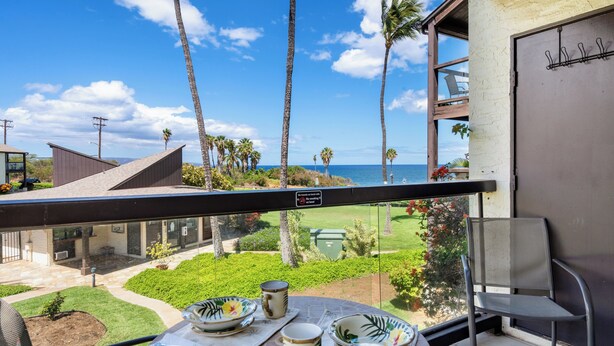 A balcony with a table set for two, overlooking a well-maintained garden and the ocean. - Hale Kamaole 208 - Relax and Enjoy Your Beautiful Ocean View Suite (Kihei)
