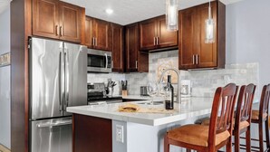 A modern kitchen with stainless steel appliances, wooden cabinets, a marble countertop, and bar stools. - Hale Kamaole 208 - Relax and Enjoy Your Beautiful Ocean View Suite (Kihei)
