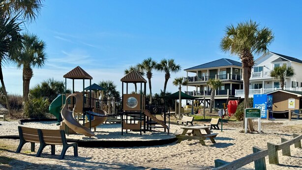A beach playground with slides and climbing structures, surrounded by palm trees and a beach house with multiple balconies. -  Ocean Lakes. Pet friendly One Story  with ramp. Complimentary golf cart. (Myrtle Beach)