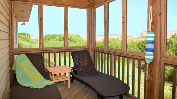 Eine überdachte Veranda mit zwei Korbsesseln, einem kleinen Holztisch und Blick auf Grünflächen und Gebäude in der Ferne.