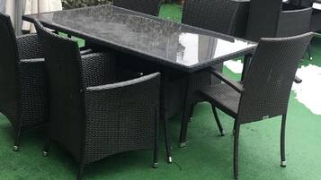 An outdoor patio with a rectangular table and six chairs, surrounded by a hedge and covered with a green artificial turf.