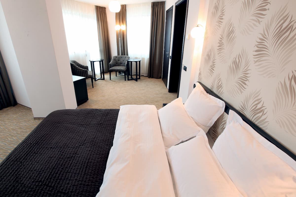 A hotel room with a large bed, a desk, and a chair. There is a window with curtains and a door in the background.