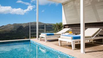 A poolside area with two lounge chairs, a small table with a drink, and a view of mountains.