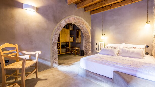 A bedroom with a bed, wooden chair, and an archway leading to a small sitting area with a table and chairs. - Rodi Hotel - Adults Only (Rhodes)