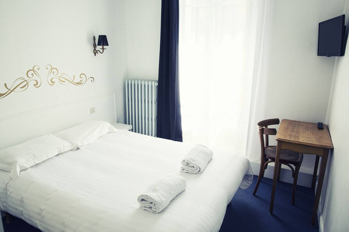 A hotel room with a bed, a chair, a desk, a TV, and a radiator.