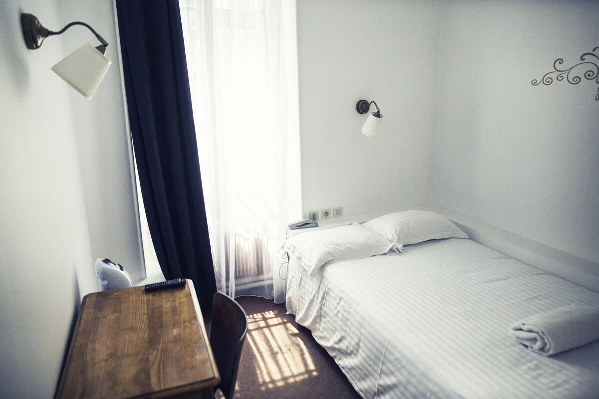 A hotel room with a single bed, a desk, a chair, a lamp, and a window with curtains.