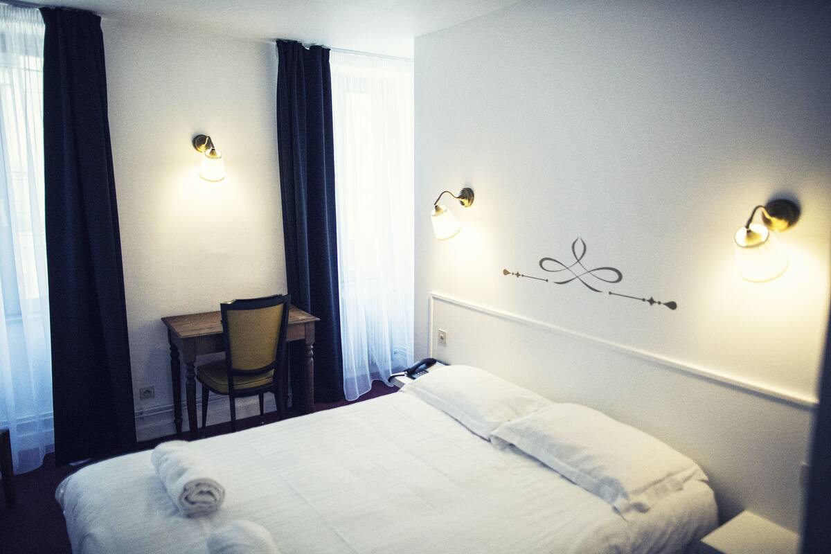A hotel room with a neatly made bed, a desk with a chair, and two wall-mounted lamps.