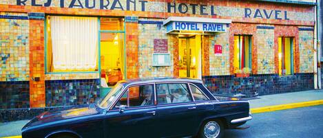 A vintage black car parked in front of a hotel with a colorful mosaic facade and a sign that reads "RESTAURANT HOTEL RAVEL".