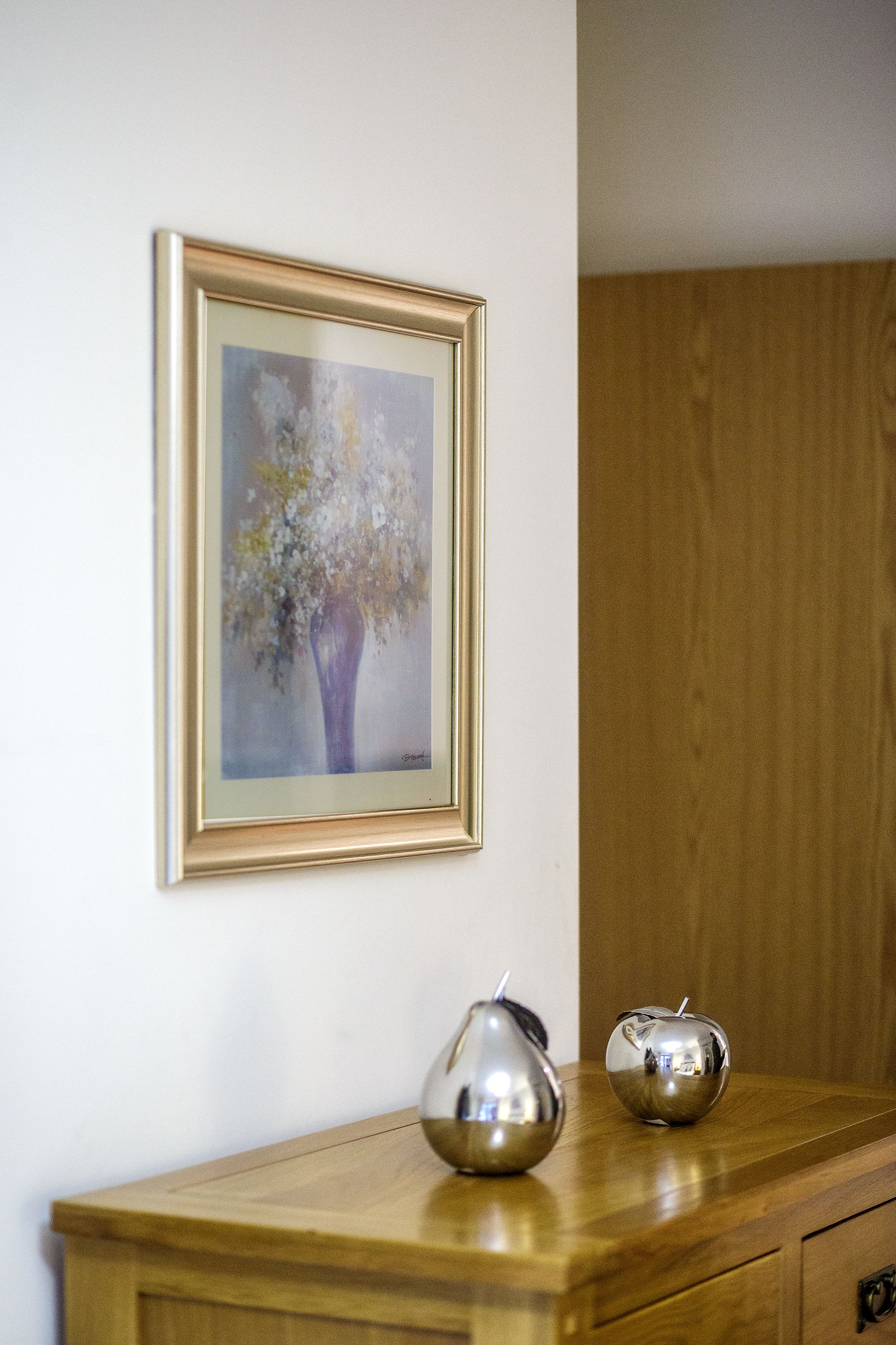 A wooden dresser with a framed painting of a vase with flowers, and two metallic vases on top.