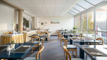 Ein Restaurant mit Tischen und Stühlen, ein Frühstücksbuffet auf der linken Seite und große Fenster auf der rechten Seite.