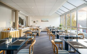 Una sala ristorante con tavoli e sedie disposti per consumare i pasti, un buffet per la colazione a sinistra e ampie finestre a destra.