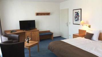 Ein Hotelzimmer mit Bett, Schreibtisch mit Fernseher, Stuhl, kleinem Tisch mit Glas und Wandregal.