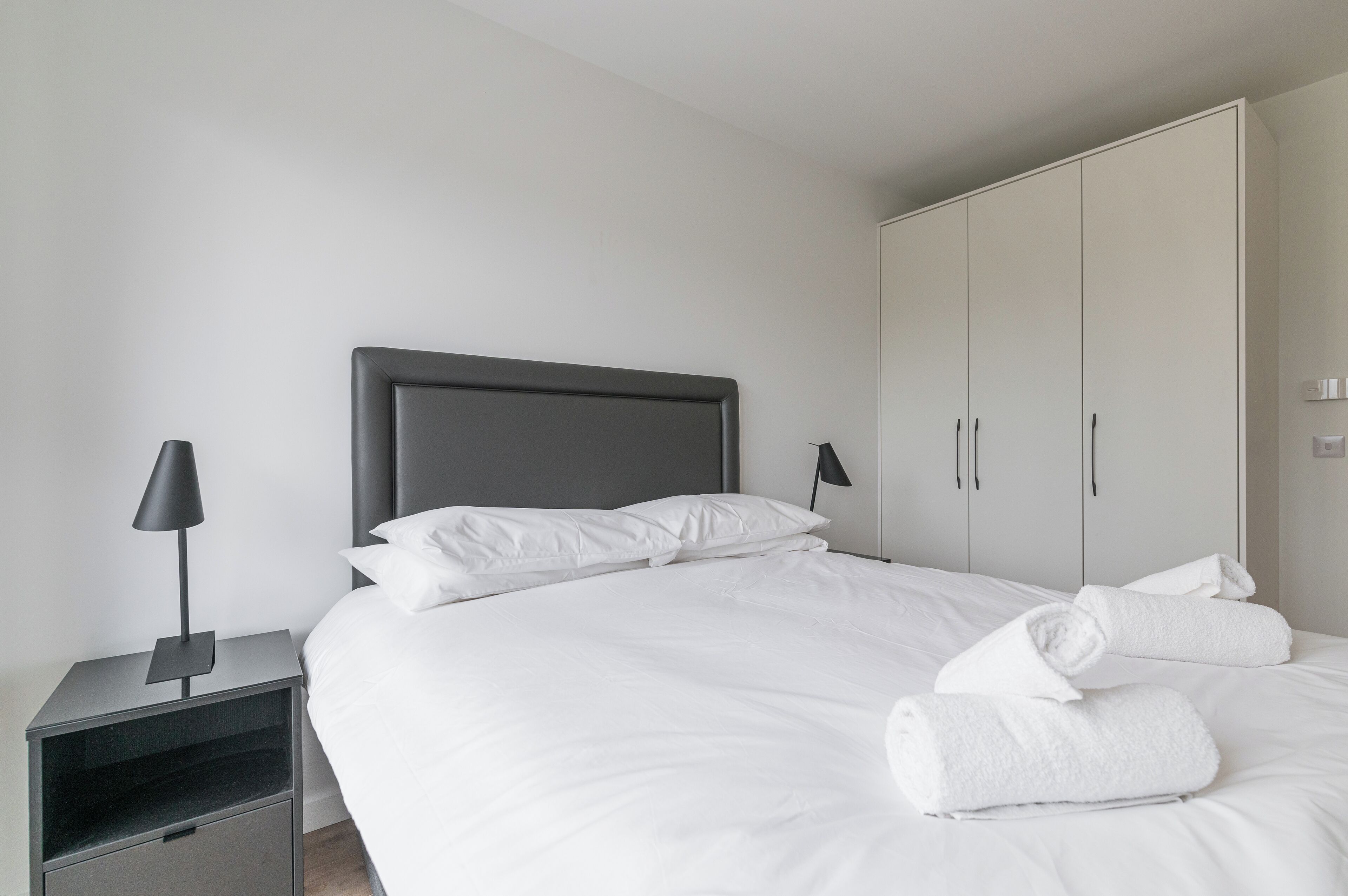 Ein ordentlich bezogenes Bett mit weißen Leinen und zwei aufgerollten Handtüchern, ein Nachttisch mit Lampe sowie ein Kleiderschrank mit Schiebetüren.
