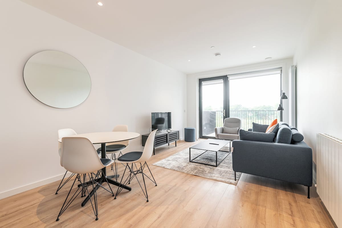 A modern living room with a round mirror, a dining area with a table and chairs, a grey sofa, and a TV.