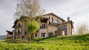 Un edificio grande de varios pisos con balcones, ubicado en una colina cubierta de césped con un árbol al frente.