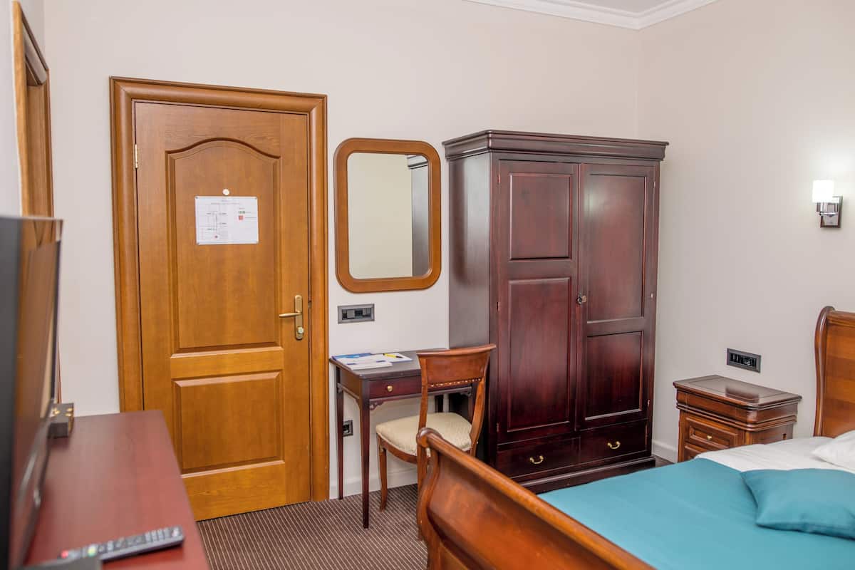 Una habitación de hotel con una puerta de madera, un escritorio con silla, un armario, una mesita de noche y una cama con colcha azul.