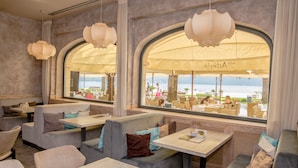 Un área de estar interior con bancos acolchados y mesas, ventanas amplias con arcos y lámparas colgantes.