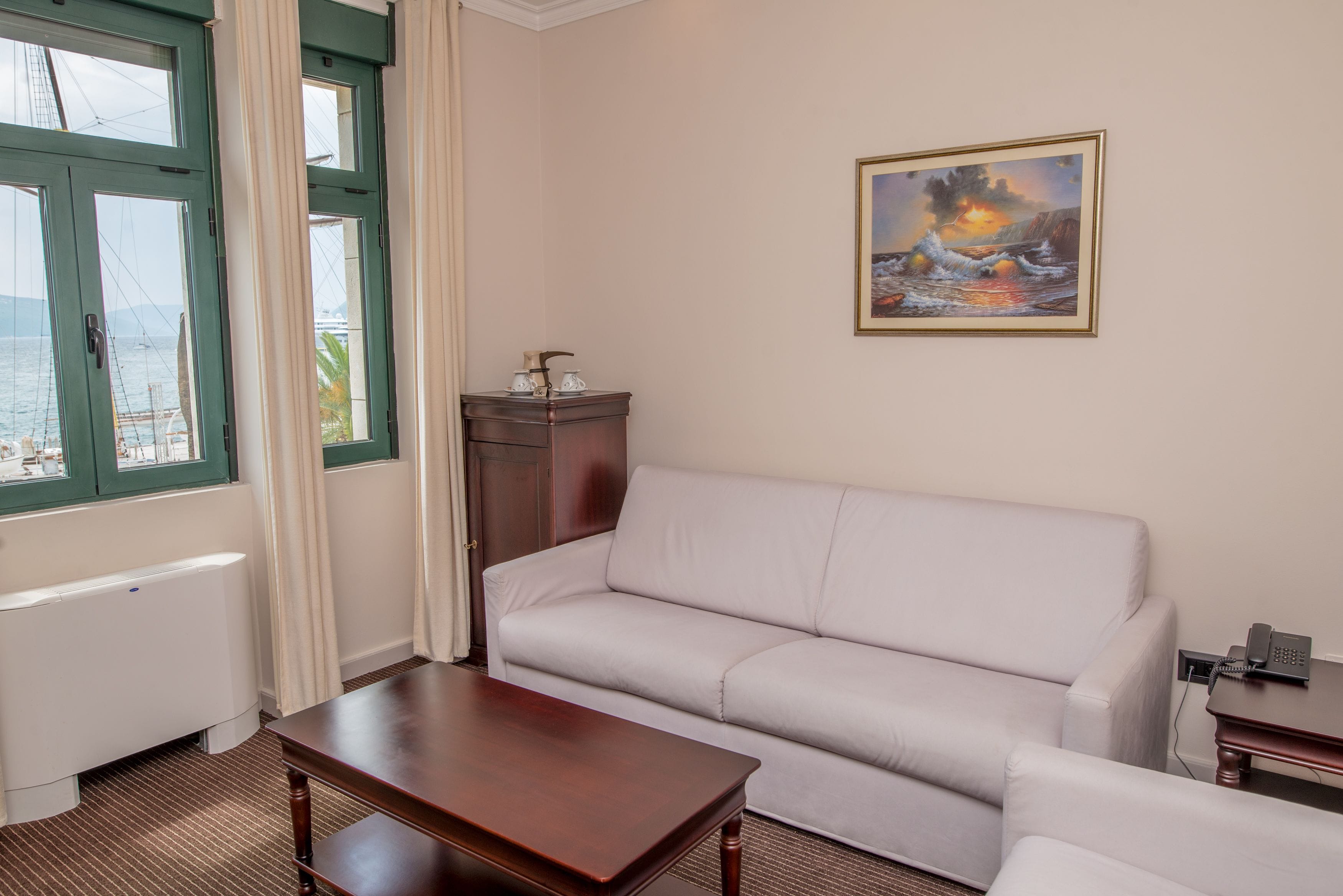 A room with a white sofa, a wooden coffee table, a painting of a sunset over the sea, and a view of the sea through a window with green frames.