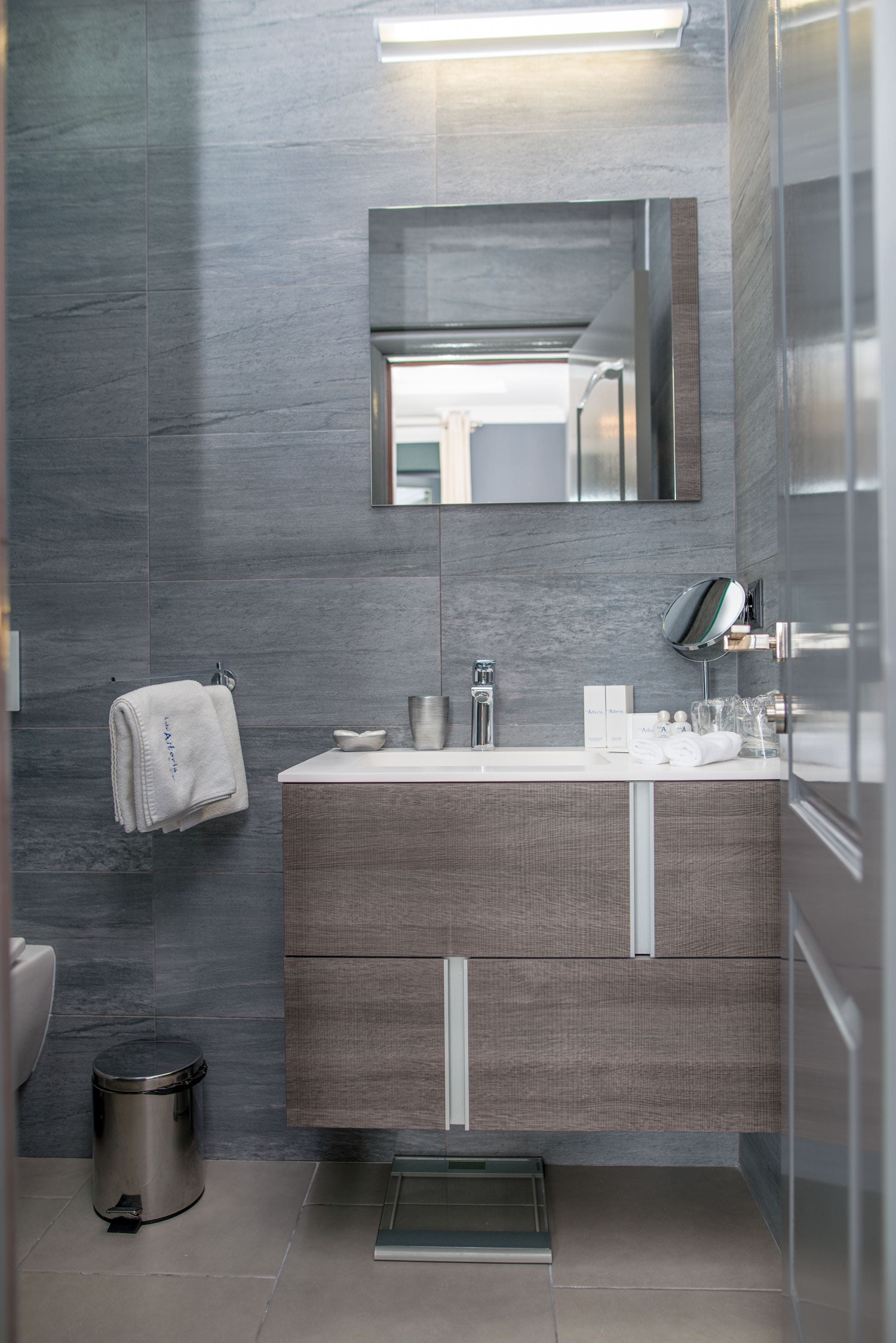 Un baño moderno con pared de azulejos grises, un lavamanos blanco con espejo superior y un pequeño cubo de basura a la izquierda.
