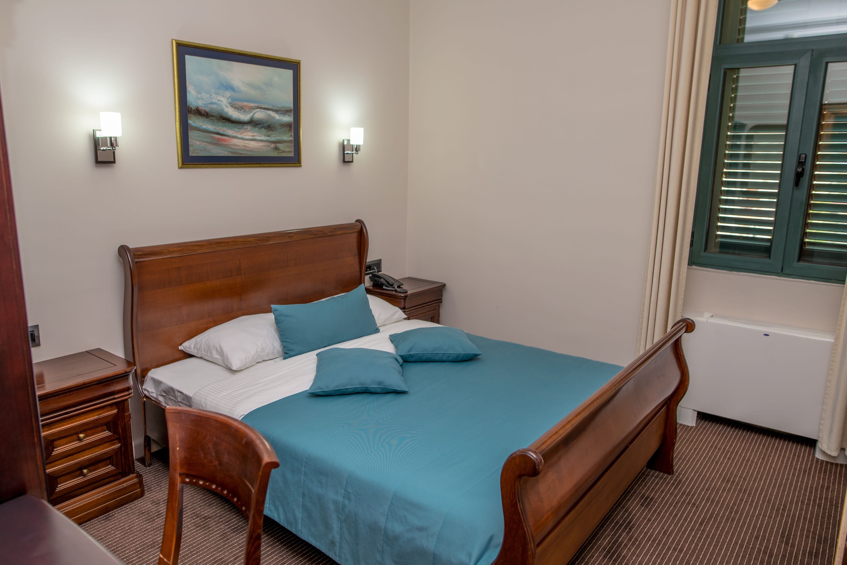 A neatly made bed with a wooden headboard, two bedside tables with lamps, a framed picture on the wall, and a window with blinds.